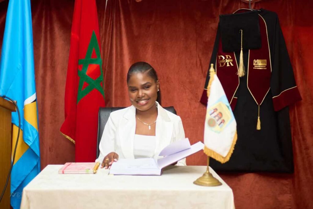 Camille Andrew assise à son bureau lors de la soutenance de sa thèse de médecine, avec les drapeaux de Sainte-Lucie et du Maroc en arrière-plan