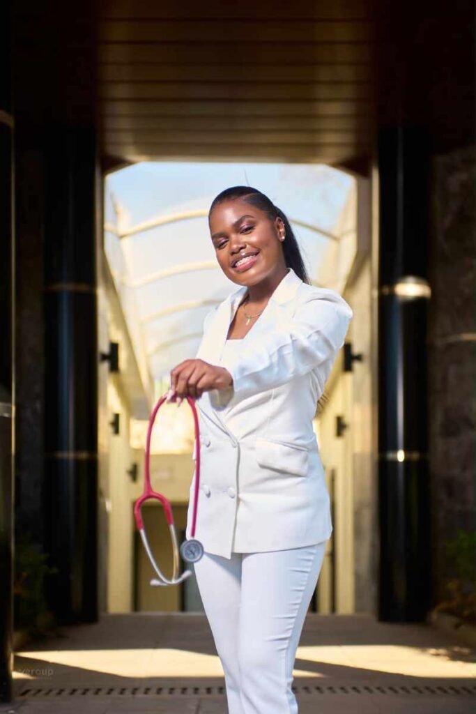 Camille Andrew en tenue blanche de médecin tenant un stéthoscope, souriante devant un bâtiment médical