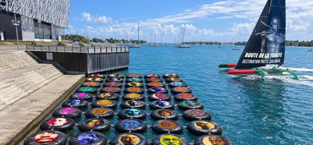 Visuel montrant des bouées ornées de portraits devant le Mémorial ACTe et un trimaran à la voile noire, en lien avec la Route du Rhum et la Guadeloupe