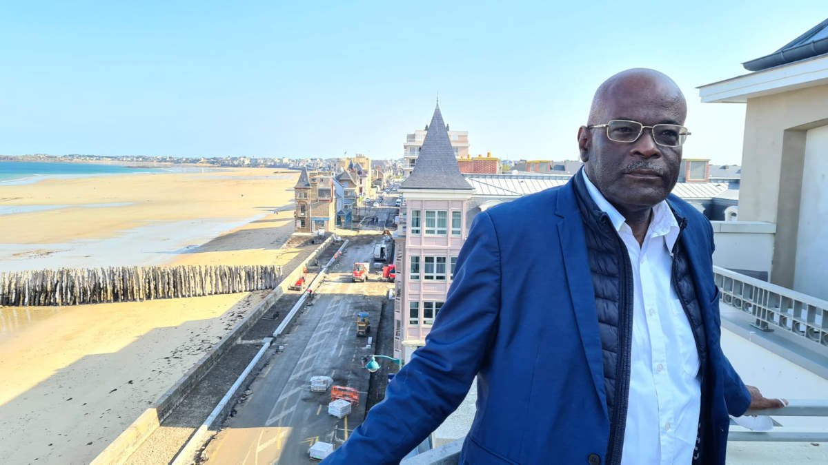 Jean-Claude Nelson en veste bleue sur un balcon, face à la plage et aux villas de bord de mer de Saint-Malo, lors de sa visite pour Caribulles